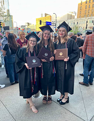Three young women in black graduation regalia holding diplomas on a city sidewalk at dusk.