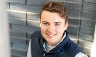 Max Milbrandt '26 smiles up at the camera against a gray brick background. He is wearing a vest with the Aldi logo.