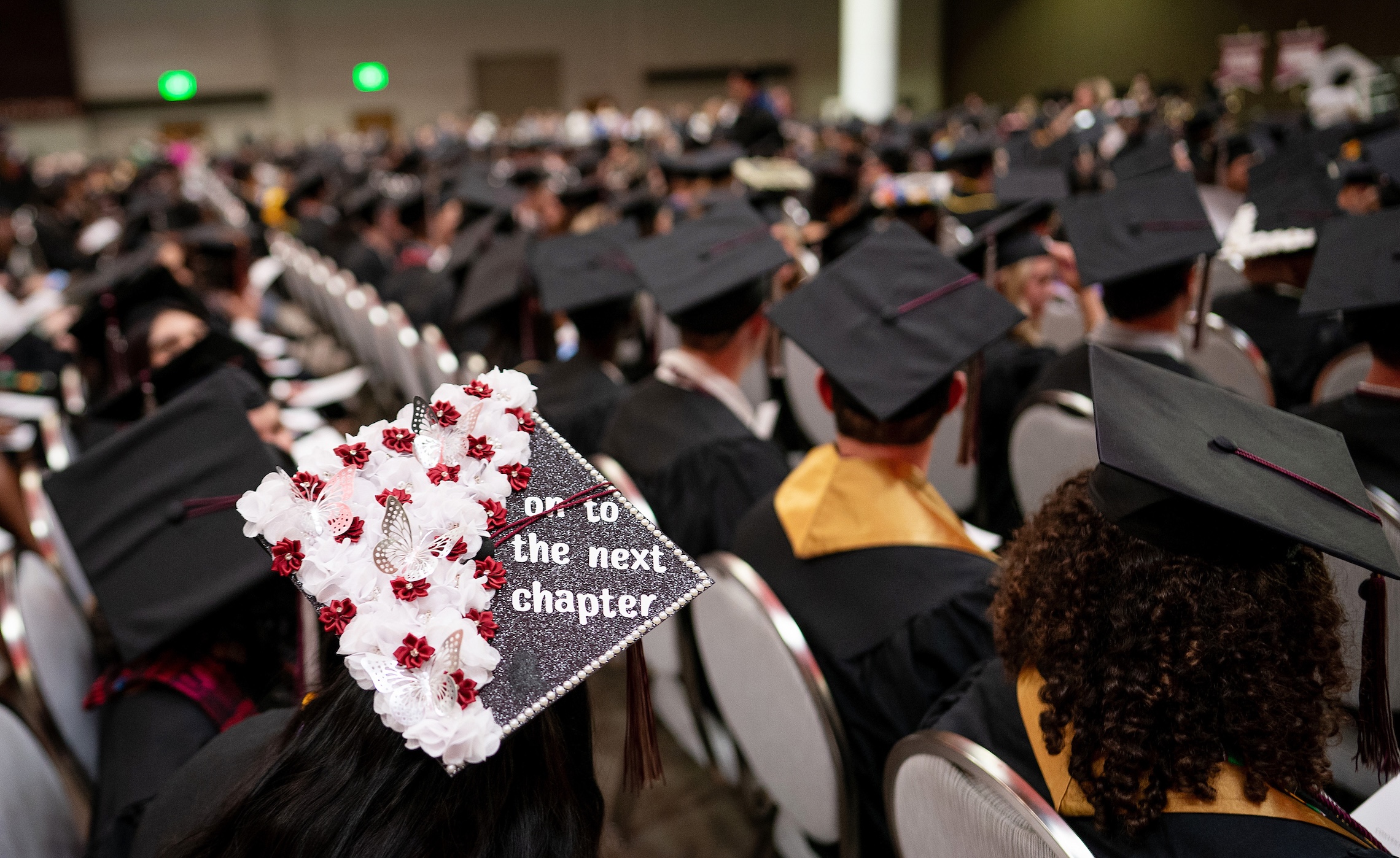 A sea of graduation hats. The closest one to the camera is decorated with red and white flowers, butterflies, and the words, "On to the next chapter."