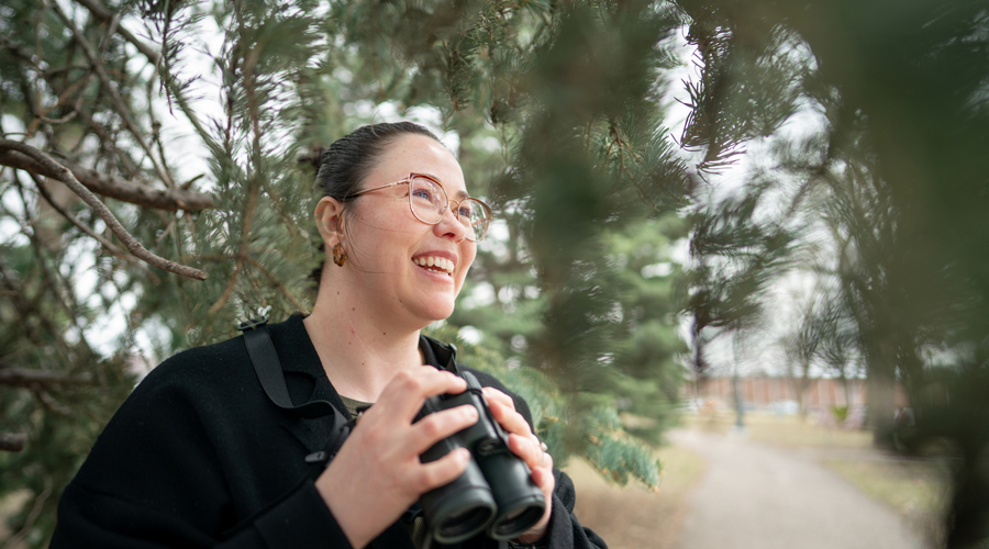 Madeline Aberg smiling while holding binoculars, standing outdoors near pine trees and a walking path.