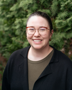 A portrait of Madeline Aberg smiling in front of a green hedge, wearing glasses and a black cardigan.