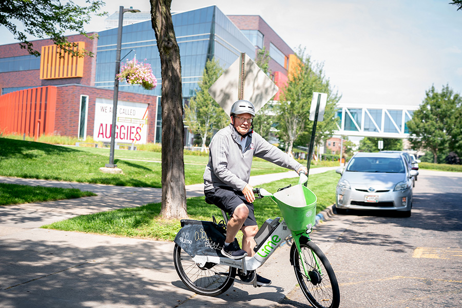 An older man wearing a helmet and a grey quarter-zip sweater rides a Lime electric bike on a paved path next to a modern brick building with a sign that reads "We are called Auggies."