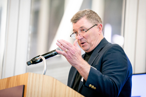 A close-up of a man with glasses speaking into a microphone at a wooden podium.