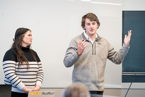 A male student gestures while speaking during a presentation as a female student in a striped sweater listens beside him.