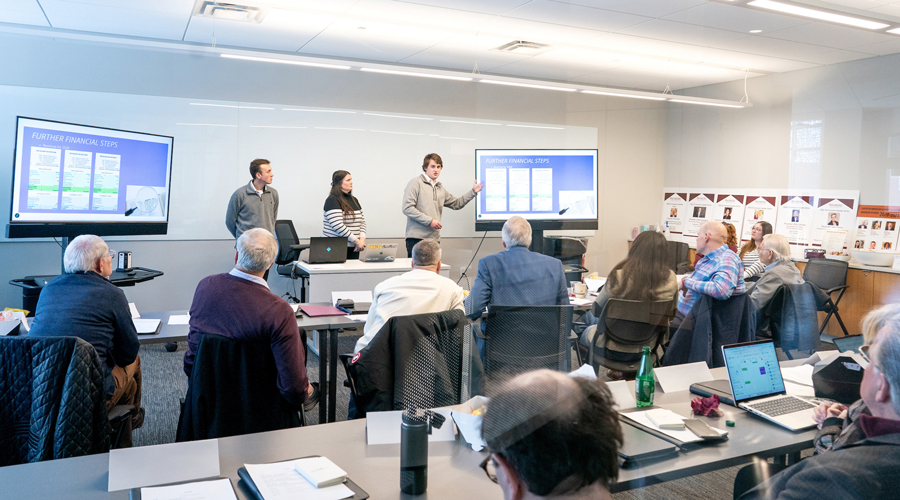 A wide shot of students presenting "Further Financial Steps" on dual monitors to a panel of judges in a bright conference room.