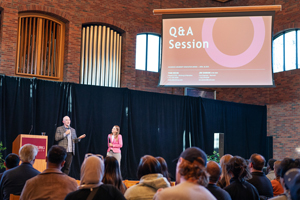 Two speakers on a stage in a large brick hall with a projection screen displaying "Q&A Session" above an audience.