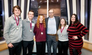 A group of four students wearing medals standing with a professionally dressed man in a modern glass-walled room at night.
