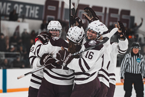 A group of Augsburg hockey players in white and maroon jerseys huddle and celebrate on the ice.