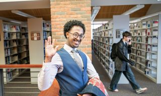 The image shows a young man in a library, standing near a wooden railing with a brick wall in the background. He has short curly hair, wears glasses, and is smiling while raising his right hand in a friendly wave. He is dressed in a blue vest over a patterned shirt with a tie. A jacket with a red patterned fabric is draped over the railing beside him. In the background, a person wearing a black jacket and jeans is walking through an aisle of shelves filled with books, holding a phone to their ear.


