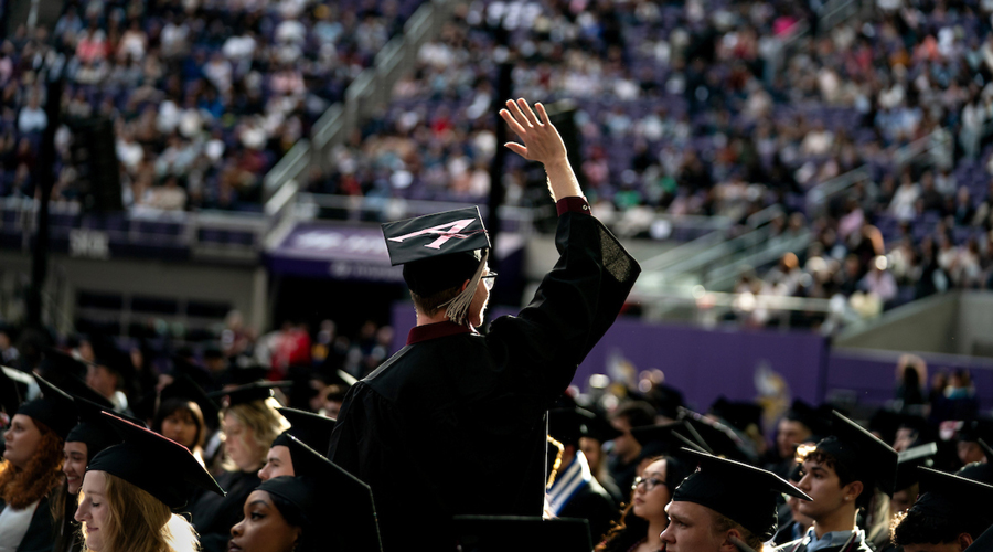 A graduate in a black cap and gown waves to a crowded stadium during a commencement ceremony.