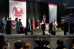 A wide shot of the commencement stage at Augsburg University, featuring faculty in academic regalia, a large university seal, and the Norwegian flag.
