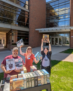 Three smiling library staff members standing outside the James G. Lindell Library entrance, holding up board games next to a cart full of games.