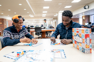 Two students smiling and playing the board game Azul inside a spacious library.