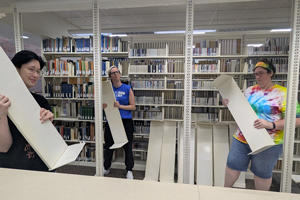 Three library staff members playfully posing while holding detached metal book shelves in front of a row of bookshelves.