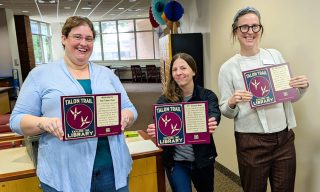 Three smiling library staff members standing inside the library, each holding up a maroon "Talon Trail" directional sign.