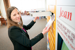 A smiling woman placing a book into an open drawer of a bright yellow automated pick-up and drop-off locker system labeled "Jean."
