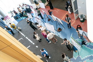 High-angle view of a poster presentation event inside a library atrium, with students and faculty gathered around trifold display boards.