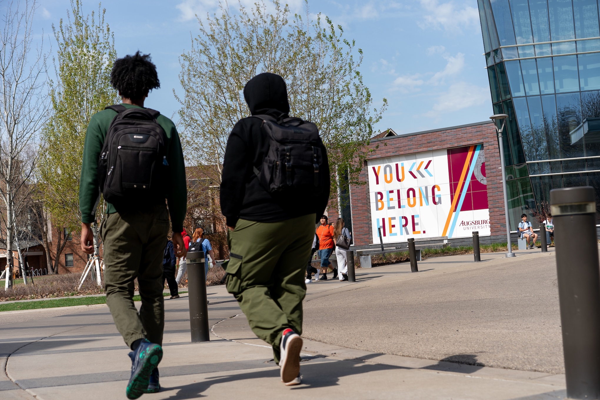 Two students wearing backpacks walk away from the camera in front of the Hagfors wall mural reading "You Belong Here"