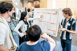 A group of three students and a faculty member gathered around a research poster titled "How Does Particle Composition Affect Organic Aerosol?" One student points to a data chart while explaining the findings.