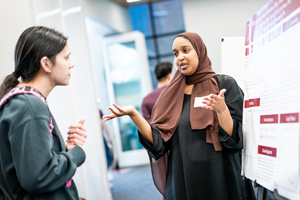 A student wearing a brown hijab uses expressive hand gestures while presenting her research poster to another student during a busy academic fair.