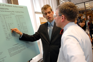 An archival photo from 2008 showing a student in a suit pointing to a data table on a research poster titled "Student Achievement in Minnesota" while talking to an evaluator.