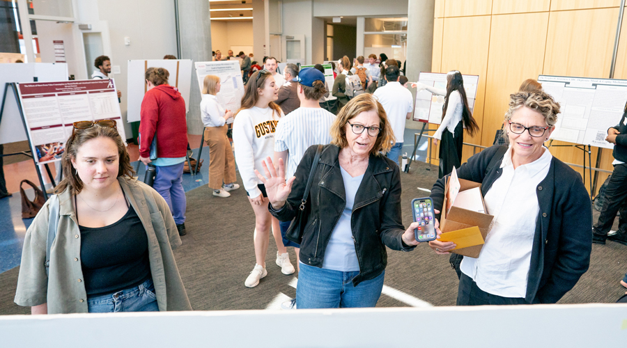 A wide shot of a bustling student research symposium in a modern campus building, with various students standing by their poster boards and attendees circulating through the hall.