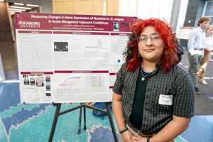 A student with bright red hair, Luis Millan, stands proudly next to his research poster titled "Measuring Changes in Gene Expression of Natalisin in D. magna."