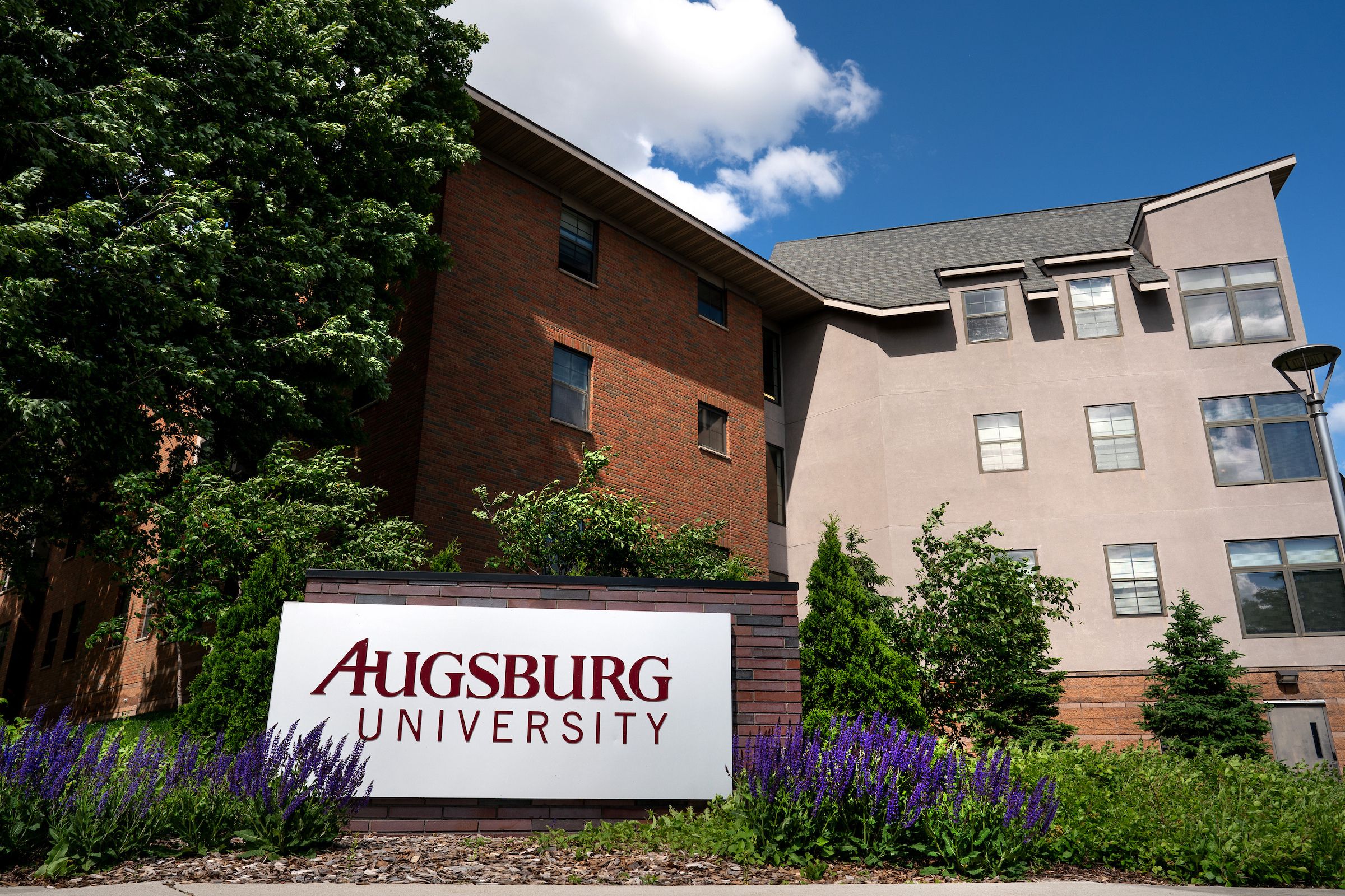 Exterior of Luther Hall with the Augsburg sign surrounded by flowers on a bright summer day.