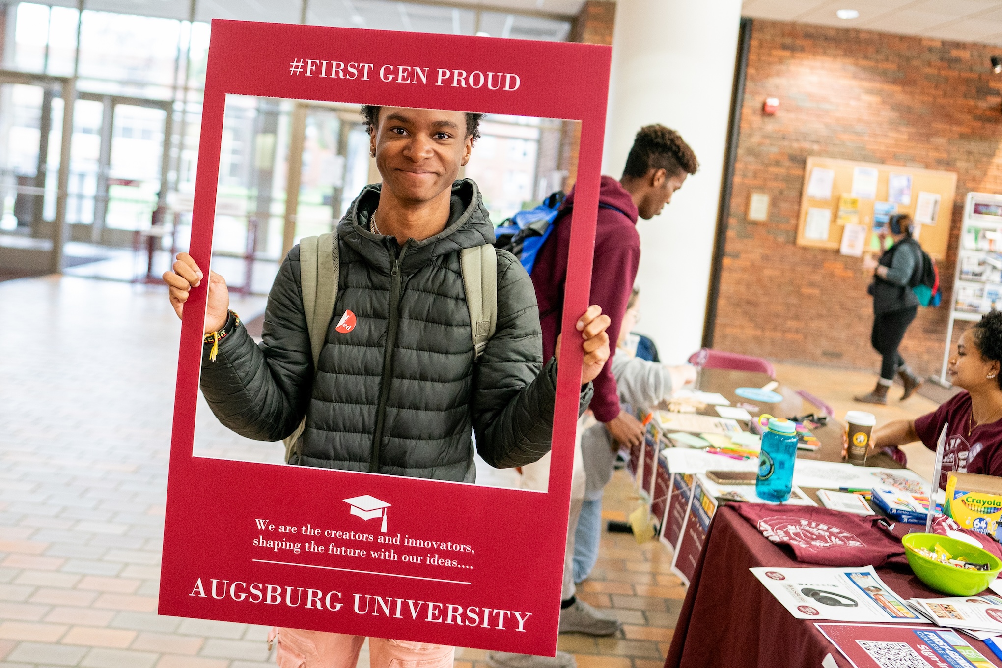 A student wearing a black puffer jacket holds up an oversized maroon photo frame around his face while standing in the lobby of Christensen Center. The frame reads "First Gen Proud."