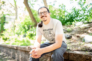 Smiling man with glasses and long dark hair tied back, wearing a graphic t-shirt and sitting outdoors on a stone wall surrounded by greenery.