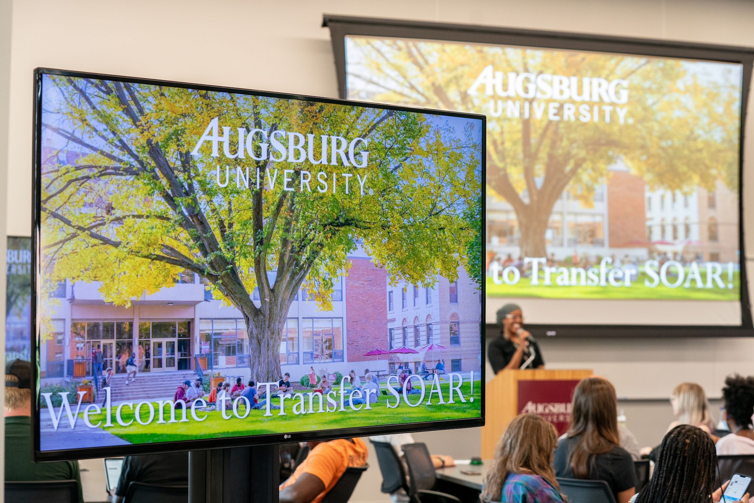 The image depicts a classroom setting at Augsburg University during a Transfer SOAR (Student Orientation, Advising, and Registration) event. In the foreground, a modern display screen shows a vibrant image of a large tree with golden leaves on the campus, with the text "Augsburg University" prominently displayed above and "Welcome to Transfer SOAR!" below. In the background, another screen displays a similar image of the tree and text. The room is filled with students seated at tables, listening to a speaker at a podium, who is partially visible on the right side of the image. The overall atmosphere suggests a welcoming and informative event for new transfer students.