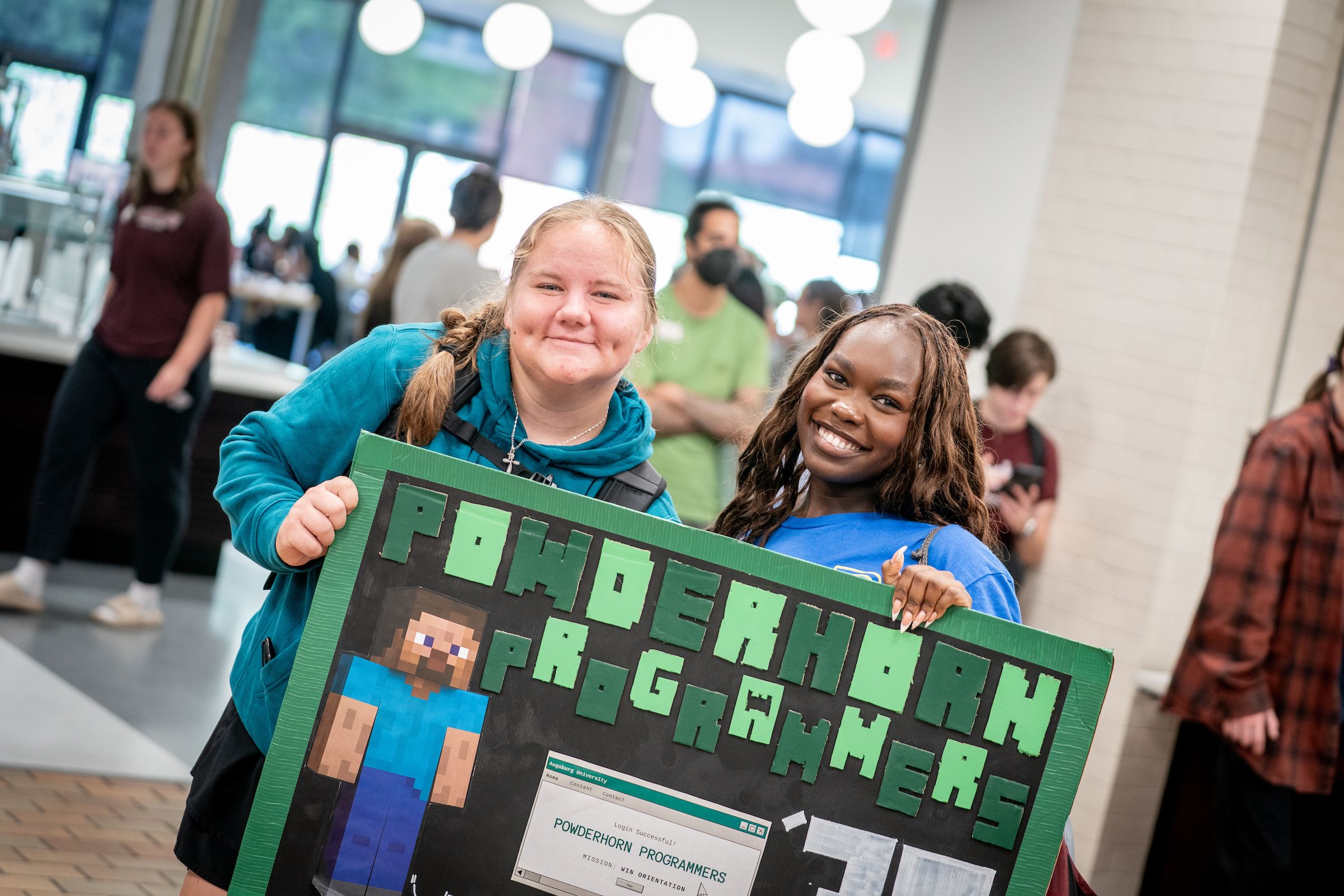 Two smiling people pose indoors while holding a large handmade poster. The person on the left wears a teal hoodie and has light hair in a side braid; the person on the right wears a bright blue shirt and has long brown hair. The poster has a green border, a black background, large raised green pixel-style lettering, and a pixel-art figure in blue clothing. A printed computer-window graphic appears near the bottom of the poster. Other people, large windows, round ceiling lights, and a tiled wall are visible in the blurred background.