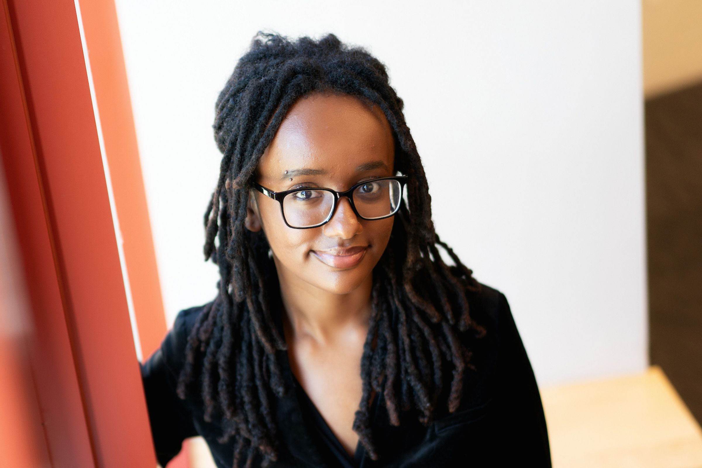A close-up portrait of a woman with long, dark hair and black rectangular eyeglasses. She faces the camera and gives a slight smile while wearing a black top. The image is brightly lit, with a white wall behind her, a reddish-orange vertical surface along the left edge, and a blurred tan surface in the lower-right corner.