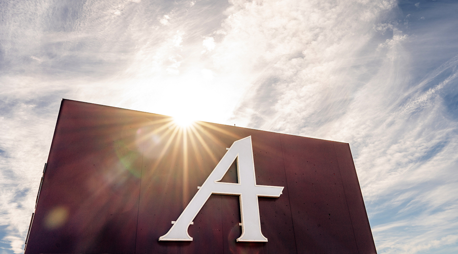 A stylized white letter "A" on a dark red sign, with sunlight bursting over the top edge against a bright, wispy blue sky.