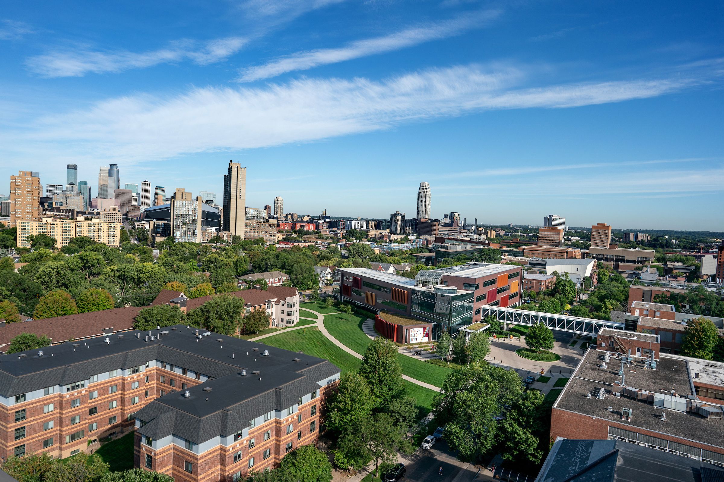A wide aerial view of an urban campus and surrounding city under a bright blue sky streaked with thin, wispy clouds. In the foreground are large brick buildings with dark roofs, tree-lined walkways, lawns, and a modern multistory building with red, orange, gray, and glass facades. A glass-enclosed pedestrian bridge crosses above a paved area near the lower right. Beyond the campus, a dense cluster of mid- and high-rise buildings stretches across the horizon, with a broad band of trees in the distance. A small sign on the modern building reads “YOU’RE BELONG HERE.”