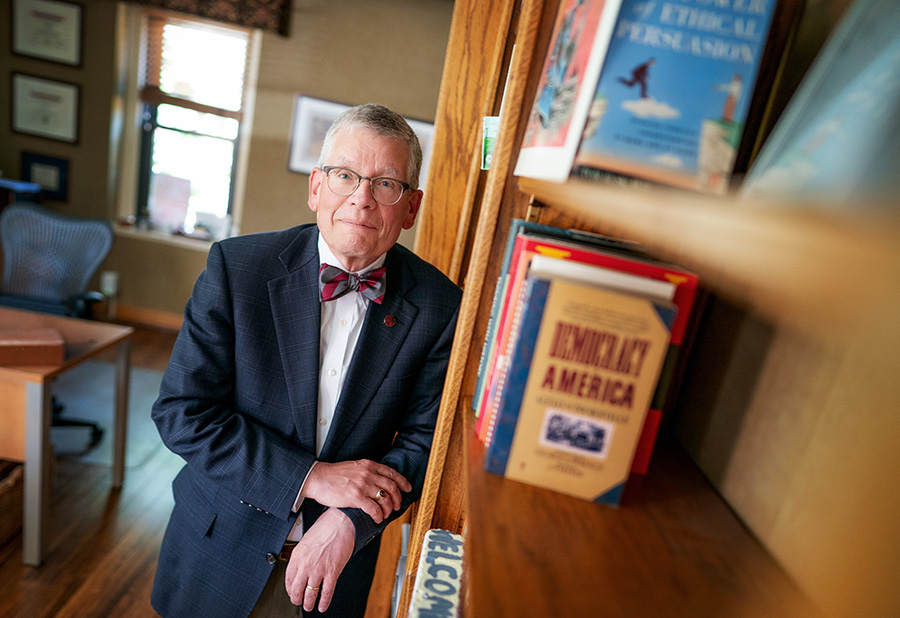 A man in glasses, a suit jacket, and a bow tie smiling next to a wooden bookshelf.