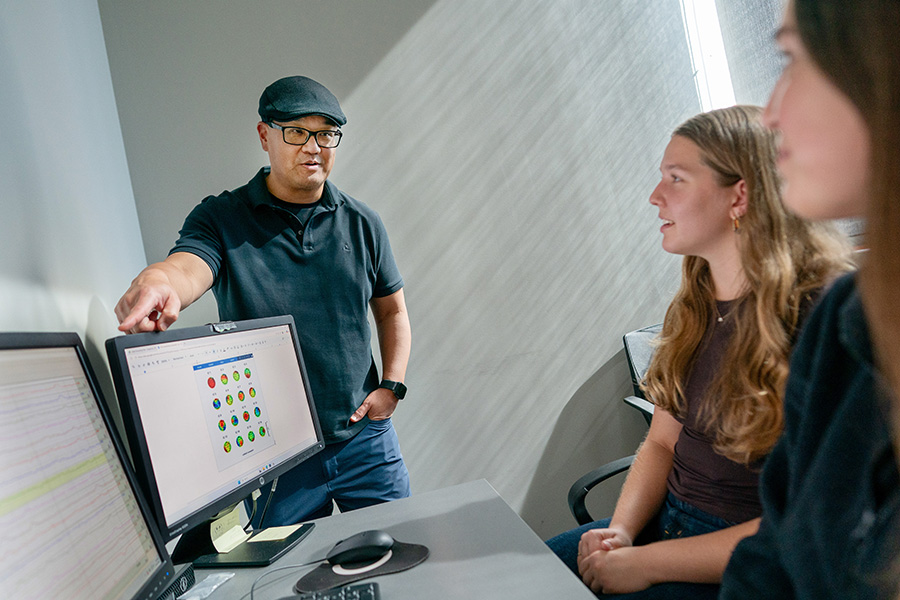 A instructor in a cap and glasses points to data on a computer screen while discussing it with two students.