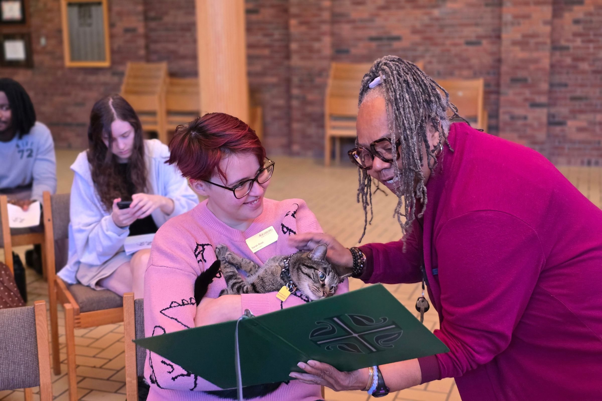 The image shows a small group gathered indoors in a brick-walled room with wooden chairs and a light-colored tiled floor. In the foreground, a person with short reddish hair and glasses, wearing a pink sweater and a name badge, cradles a gray tabby cat. An older adult with gray braids, glasses, and a burgundy top leans toward the cat while holding an open dark-green folder or book. Two other people sit in the background; one looks down at a phone, and another holds a white paper. Framed items hang on the brick wall behind them.