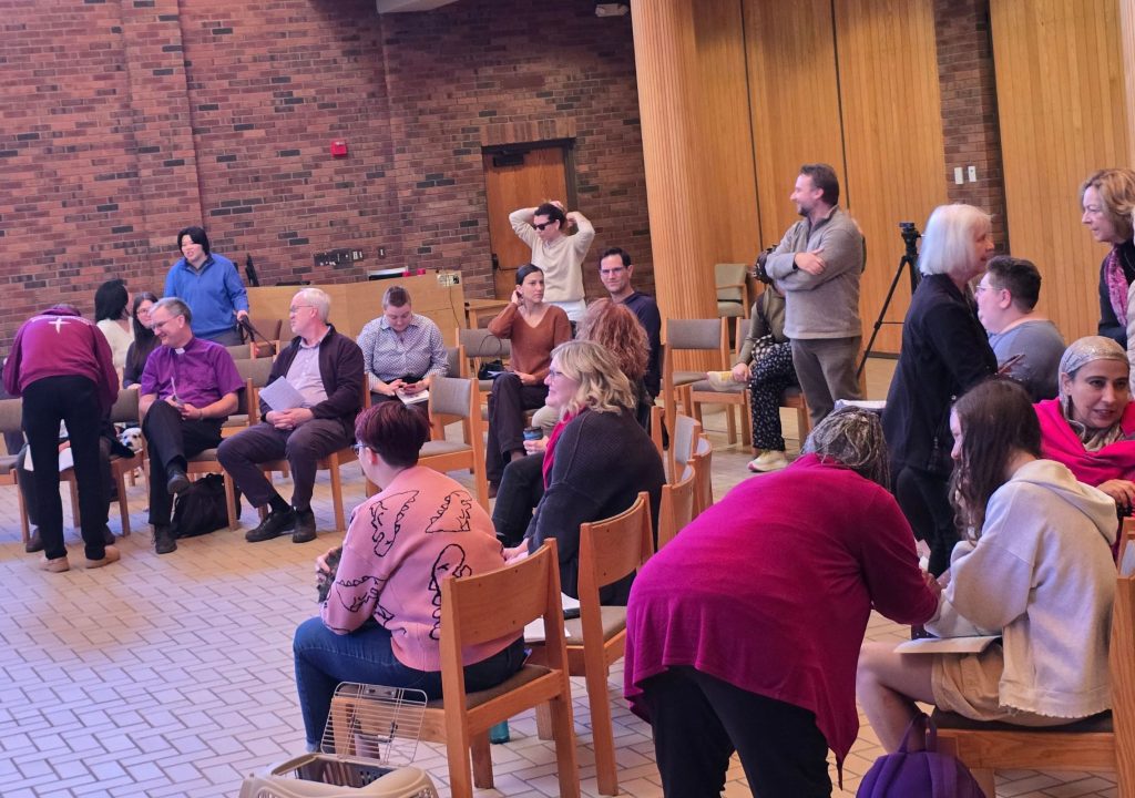 11 A group of adults gathers in a large indoor room with exposed reddish-brown brick walls, light wood paneling, and rows of wooden chairs. Some people are seated while others stand, talk, or move between the chairs. A seated person near the left wears a purple clerical shirt with a white collar. Near the center, one person holds papers, and another adjusts their hair. A camera on a tripod stands among the chairs on the right. The tiled floor, wood-paneled doors, electrical outlets, and a small red wall fixture are visible in the background. Several people in the foreground are seen from behind, including individuals wearing bright pink and magenta clothing.