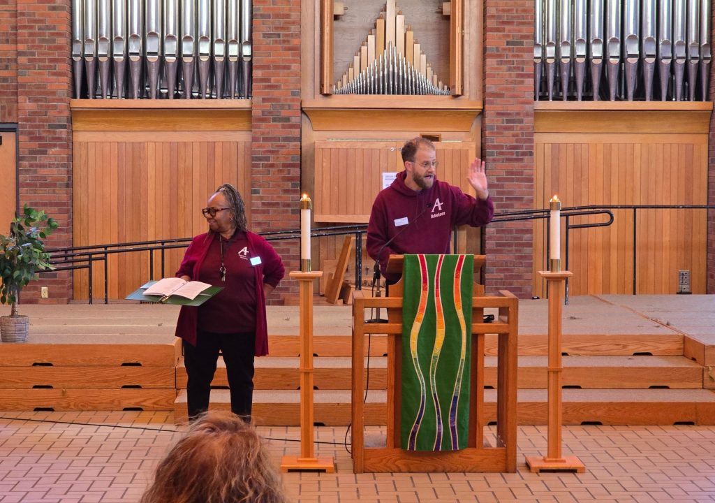 2 A church or chapel interior with a large pipe organ set into a red-brick wall. Three sections of tall silver organ pipes are visible above wooden panels, with the central section arranged in a peaked pattern. In the foreground, a man wearing glasses and a maroon sweatshirt stands behind a wooden lectern and raises one hand. The lectern is draped with a green cloth decorated with red, yellow, blue, and white curved stripes. To his left, a woman in a matching maroon top holds an open book or binder. Wooden steps, railings, candle stands, a potted plant, and tiled flooring surround them.