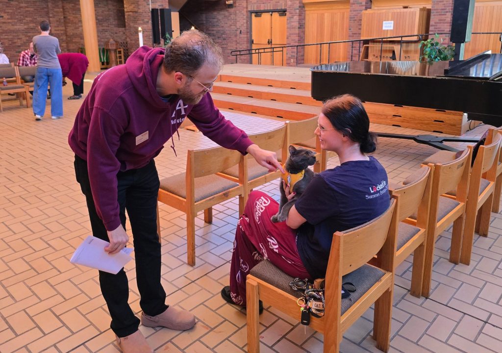 5 A group of people gathers in a brick-walled indoor space with a raised wooden platform, rows of wooden chairs, and a black grand piano. In the foreground, a man wearing a burgundy hoodie leans toward a seated student and gestures toward a gray cat they are holding. The cat wears a yellow harness and looks toward the man. The student wears a dark shirt and burgundy pants with white lettering. Several people sit or stand in the background near additional chairs. The floor is covered with light-colored rectangular tiles, and potted plants, candles, doors, and accessibility railings are visible around the room.