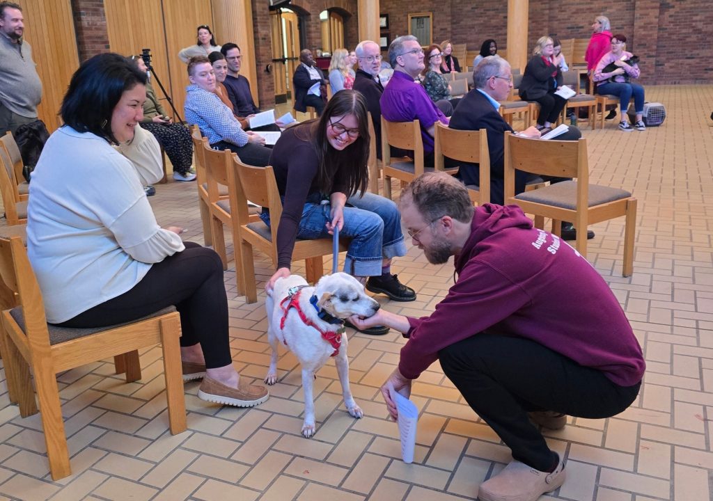 6 A group of people sits in rows of wooden chairs inside a brick-and-wood building. In the foreground, a small white-and-tan dog wearing a red harness stands between the chairs while a kneeling man in a maroon shirt pets it. A seated staff member and a smiling individual with glasses are nearby. Additional attendees, some holding papers, fill the background. The room has a tan tiled floor, brick walls, wood-paneled sections, and large wooden columns.