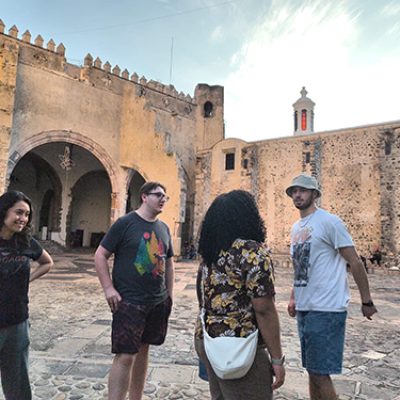 764892e4-6cf5-441f-a667-89c26b3a92d4 Four young adults gathering and talking in front of a historical stone building and courtyard.