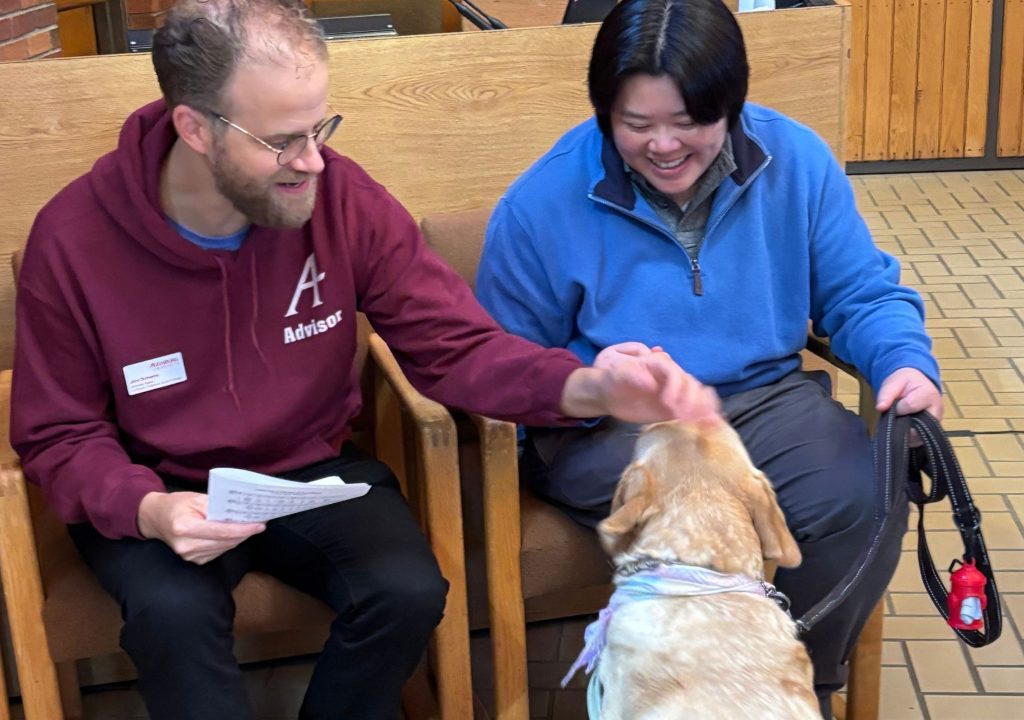 8 Two adults sit in wooden armchairs in an indoor waiting area while interacting with a light-colored dog in the foreground. The person on the left wears glasses and a burgundy hoodie labeled “A Advisor,” holds a sheet of paper, and reaches toward the dog. The person on the right wears a bright blue jacket and smiles while looking down at the dog. The dog wears a blue-purple collar or harness and faces away from the camera. Behind them are wood-paneled walls, a brick section, and a tan tiled floor.