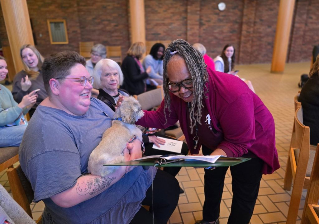 9 A group of people gathers in a brick-walled room with wooden columns and rows of chairs. In the foreground, a seated person wearing glasses and a gray shirt holds a small, fluffy cream-colored dog with a blue harness. A person with long gray braids, glasses, and a burgundy shirt leans toward them while looking at the dog. Papers and an open binder rest between them. Several people sit and talk in the background.