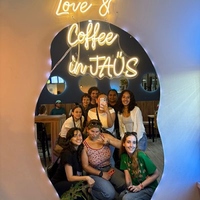 IMG_3004 Group of friends posing for a selfie reflection in a coffee shop mirror under a glowing neon sign reading "Love & Coffee in JAÜS".