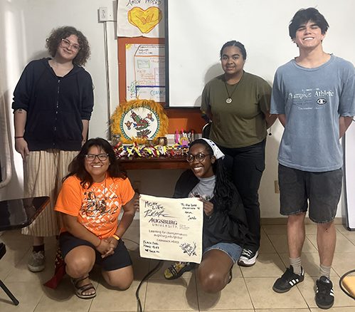 : Five students group together in a classroom, with one student in the center holding up a signed Augsburg University tote bag.