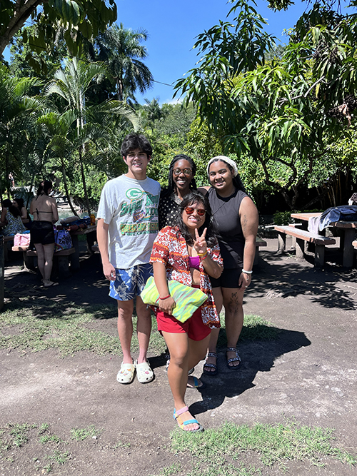 Four friends pose outdoors under lush green trees near picnic tables on a sunny day.