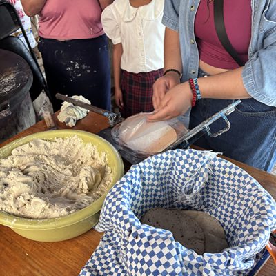 IMG_6852 2 A person presses masa dough near a bowl of dough and a basket lined with a blue-and-white checkered cloth holding fresh tortillas.