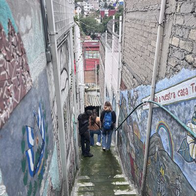 IMG_8226 2 People walk down a narrow outdoor staircase framed by high brick walls decorated with colorful painted murals.